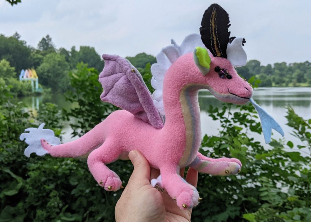 a pink dragon with a pastel stripes underbelly, aqua and yellow ears, gray and purple wings, white mane/tail/elbow tufts, gold claws/markings, and black & gold mothlike antennae and golden purple glass eyes