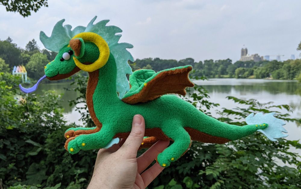 a green dragon plushie with a warm brown underbelly/underwings/inner ears, yellow horns/claws/markings, a purple tongue, aqua mane/tail/elbow tufts, and white eyes