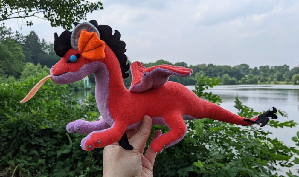 a pink-red dragon plushie with a lilac/light purple underbelly and underwings, bright orange inner ears, grey/silver horns/claws/markings, sky blue eyes, a peach tongue, and a black crest and tailtuft