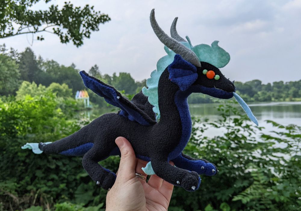 a black dragon plushie with a dark blue underbelly/underwings/inner ears, light aqua mane/tail/elbows/eye details, orange eyes, light blue tongue, and gray/silver horns/claws/markings