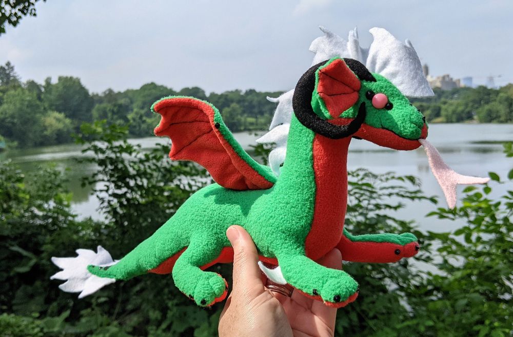 a watermelon colored dragon plushie (bright green with pink-red underbelly & accents, black claws/horns, white mane/tailtip, pink eyes)