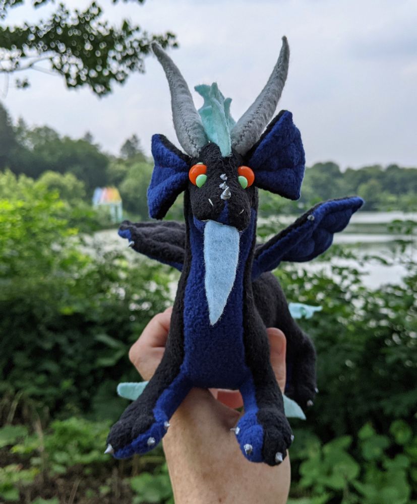 a black dragon plushie with a dark blue underbelly/underwings/inner ears, light aqua mane/tail/elbows/eye details, orange eyes, light blue tongue, and gray/silver horns/claws/markings