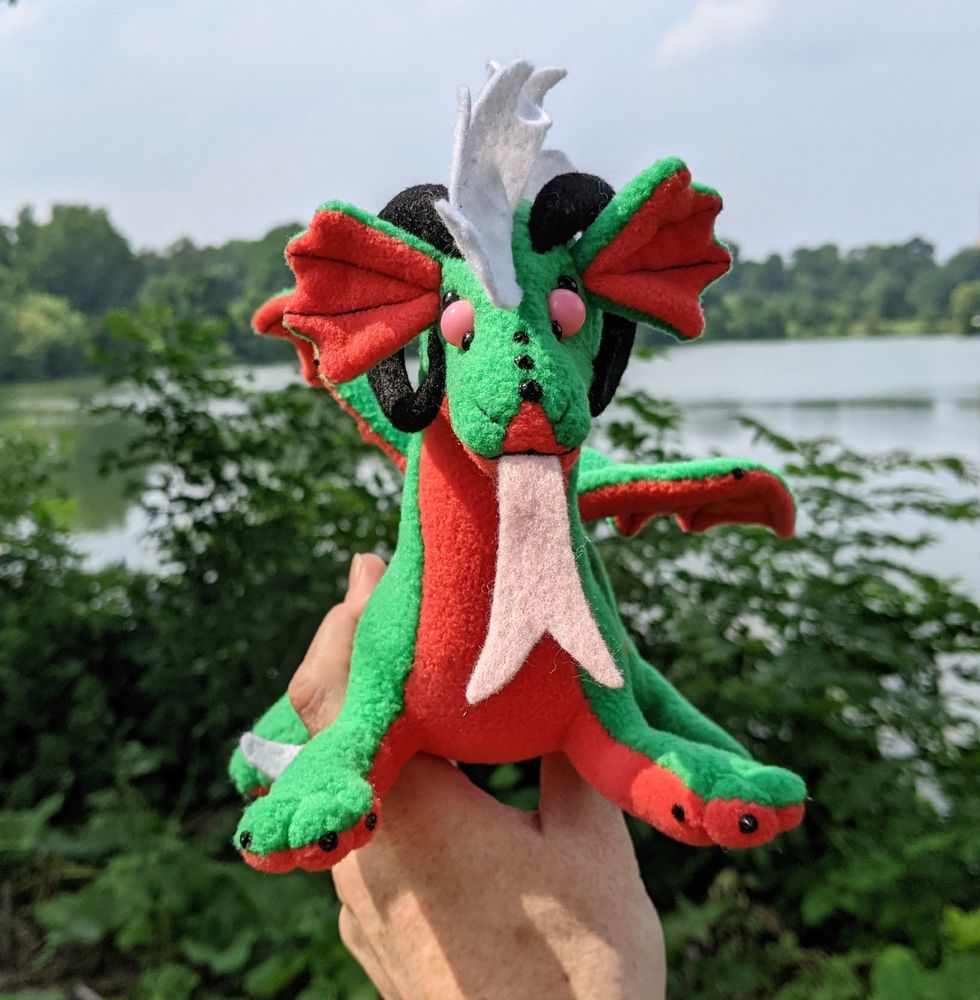 a watermelon colored dragon plushie (bright green with pink-red underbelly & accents, black claws/horns, white mane/tailtip, pink eyes)
