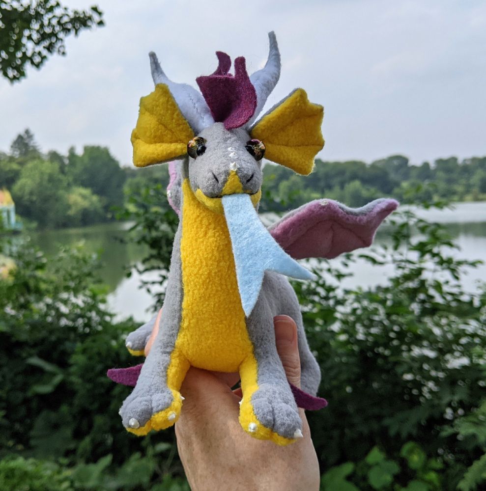 a soft gray dragon plushie with a honey yellow underbelly and inner ears, light purple/lilac underwings, raspberry colored mane/tailtuft/elbow tufts, white horns/claws/markings, and golden purple glass eyes