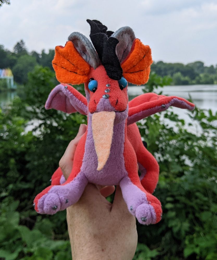 a pink-red dragon plushie with a lilac/light purple underbelly and underwings, bright orange inner ears, grey/silver horns/claws/markings, sky blue eyes, a peach tongue, and a black crest and tailtuft