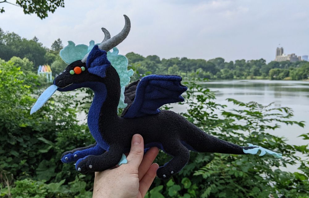 a black dragon plushie with a dark blue underbelly/underwings/inner ears, light aqua mane/tail/elbows/eye details, orange eyes, light blue tongue, and gray/silver horns/claws/markings