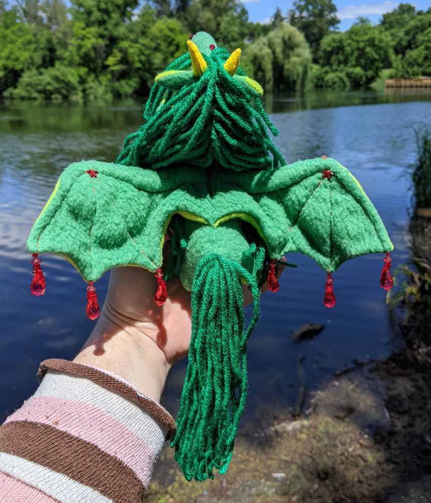 the back of a green bat-winged horse plushie that's also dragon. there's yellow horns, red translucent beads catching the light, detailing some bead work by the wing joints. they also have a green yarn mane and tail. there is a lake in the background