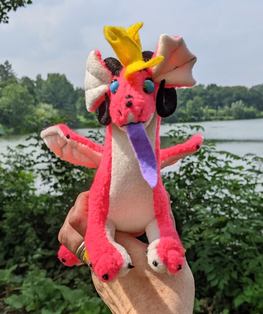 a bright neon pink dragon with a white underbelly/inner ears/underwings, purple tongue, yellow mane/tail/elbow tufts, black horns/claws/markings, sky blue eyes