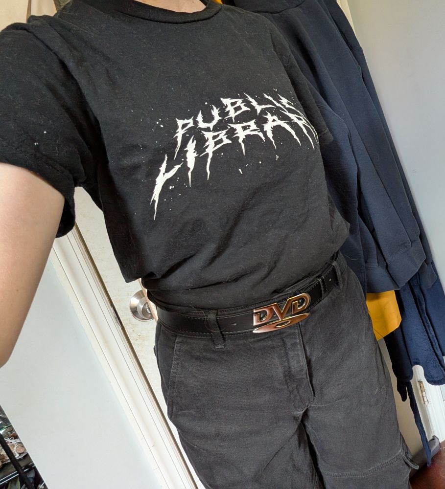 black shirt with public library written in heavy metal font, with black pants and black belt with metal DVD logo buckle