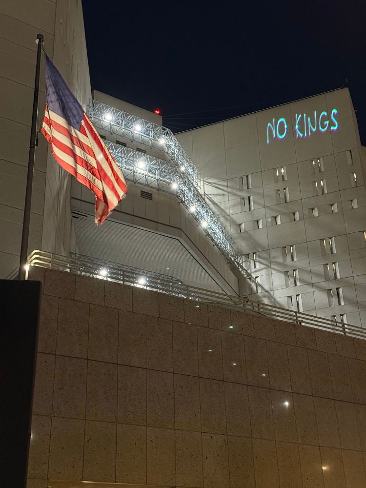 Word “No Kings” projected on a jail building while the United States flag flaps below.
