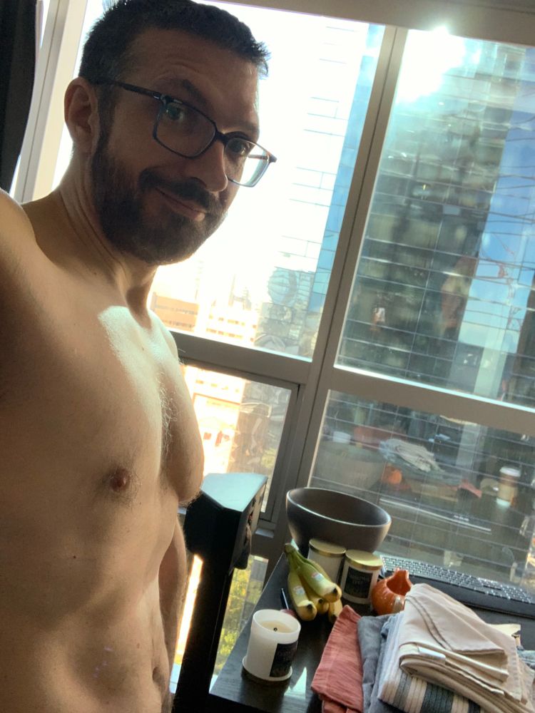 A person taking a no shirt selfie in front of a window and trying hard not to feel weird about it.
