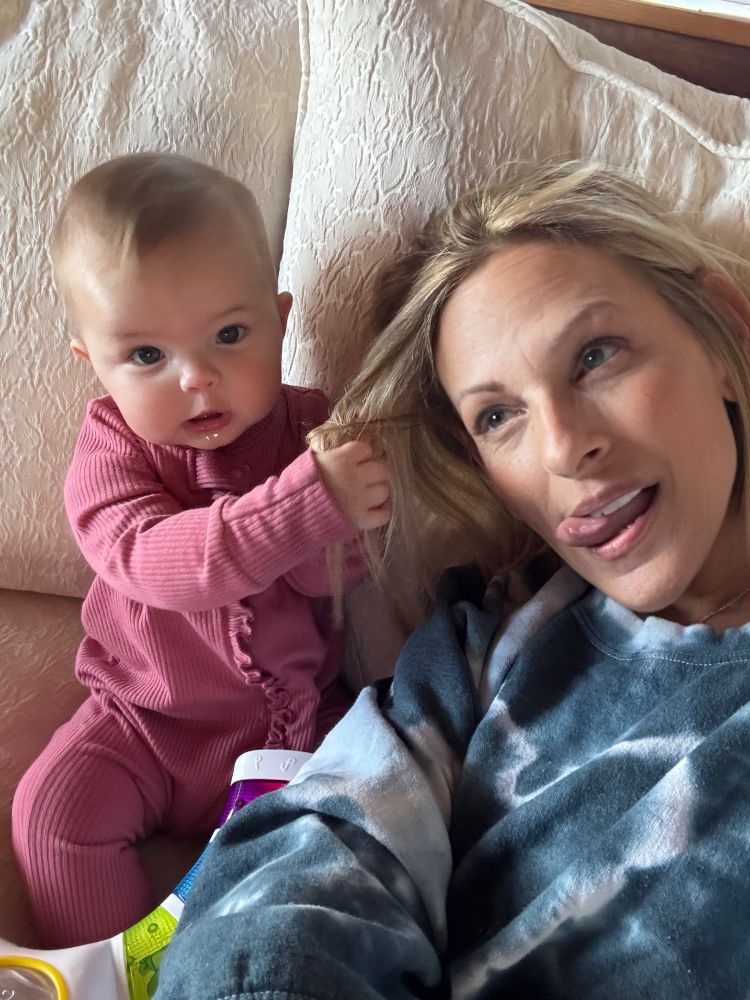Marlee Matlin is laying on a bed wearing a blue tie dyed sweatshirt while her 7 month old grandaughter in pink onesie is pulling playfully on her hair