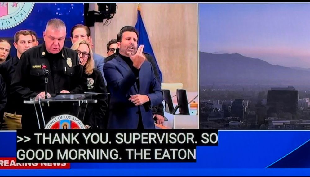 A sign language interpreter signs while standing next to the police chief of La Angeles who is speaking at a podium during a press conference. 