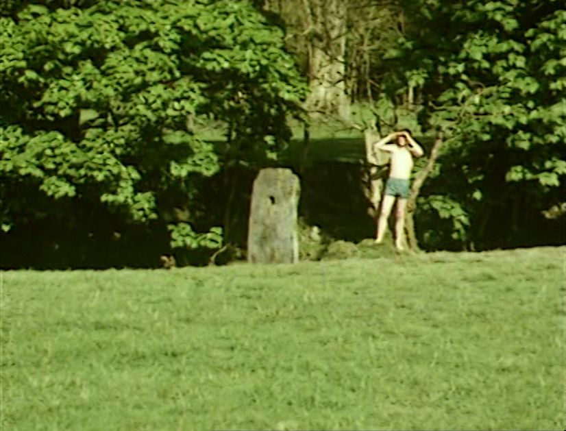 Screenshot from episode 1 of Granada TV’s The Owl Service (1969). Roger (Francis Wallis), standing on a riverbank, shields his eyes from the sun as looks across a valley. He is bare chested and wearing green shorts. A stone with a hole bored through stands nearby.