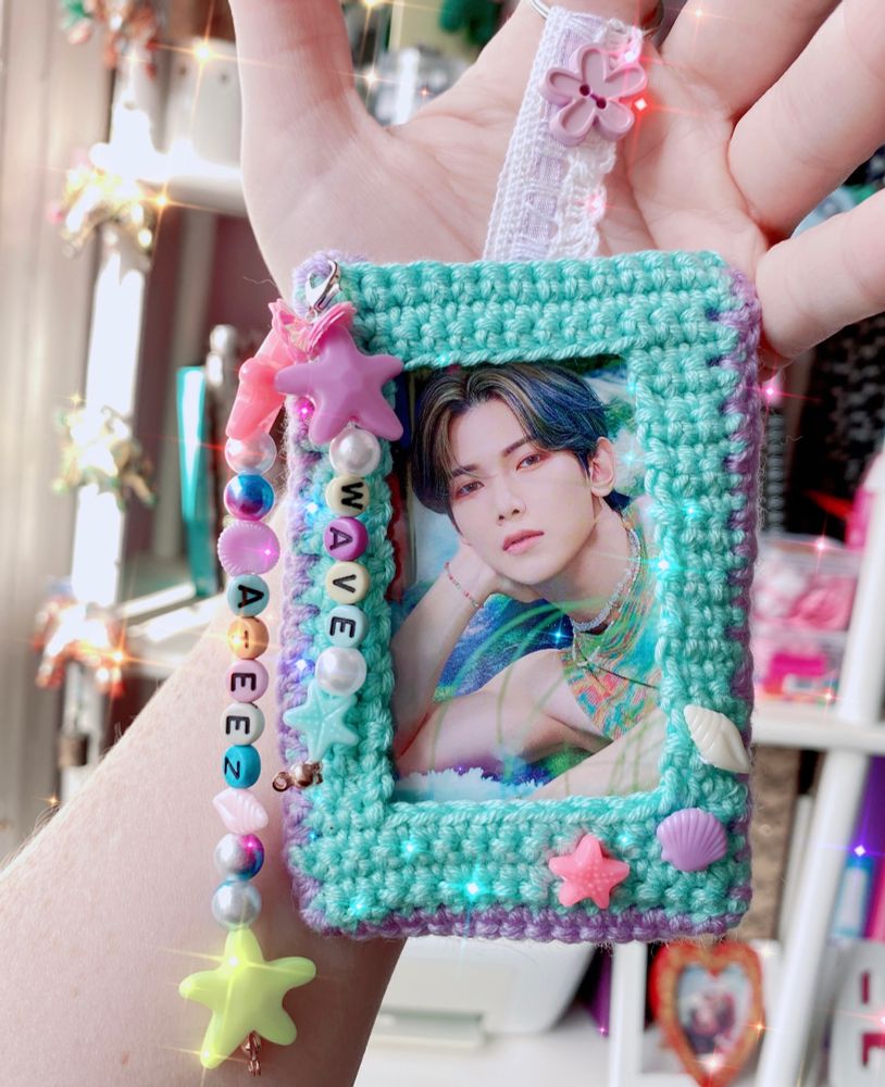 Crocheted photocard holder in aquamarine and lilac wool. There are pastel shell details and hanging beads spelling out Ateez and Wave. There is a cute photo inside of Yeosang from Ateez wearing similar colours, and there is a sparkle filter over the photo