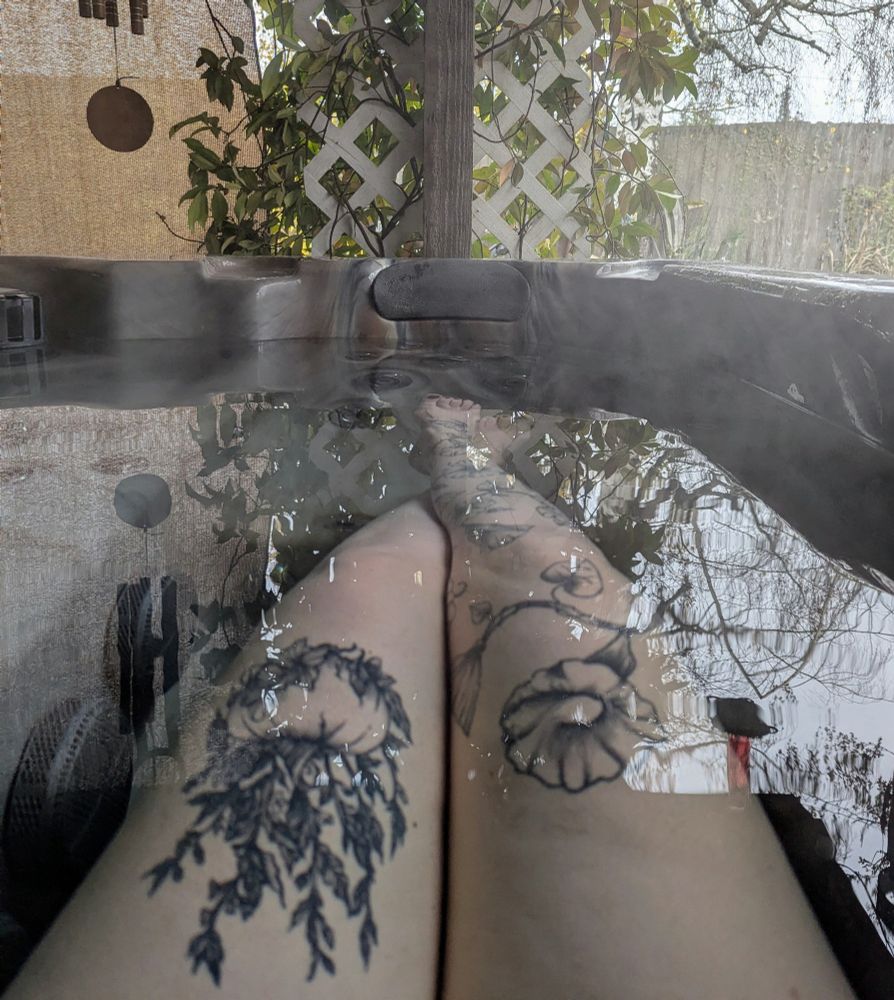 Tattooed legs in a hot tub with bare tree branches reflected on the water