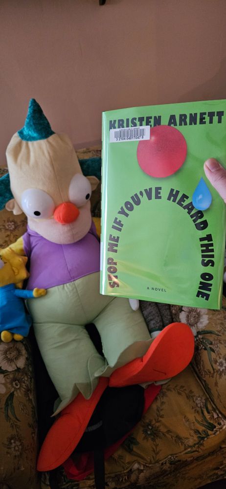 Photo of hardcover copy of "Stop Me If You've Heard This One" by Kristen Arnett, with a giant Krusty the Clown doll in the background.