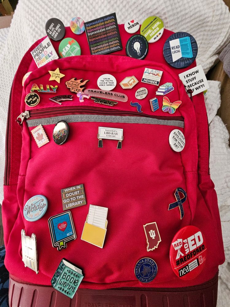 New enamel pin two hands holding up a sign that says, "This is what a LIBRARIAN looks like" from Not Yo Mama's Librarian on my red backpack with other pins.