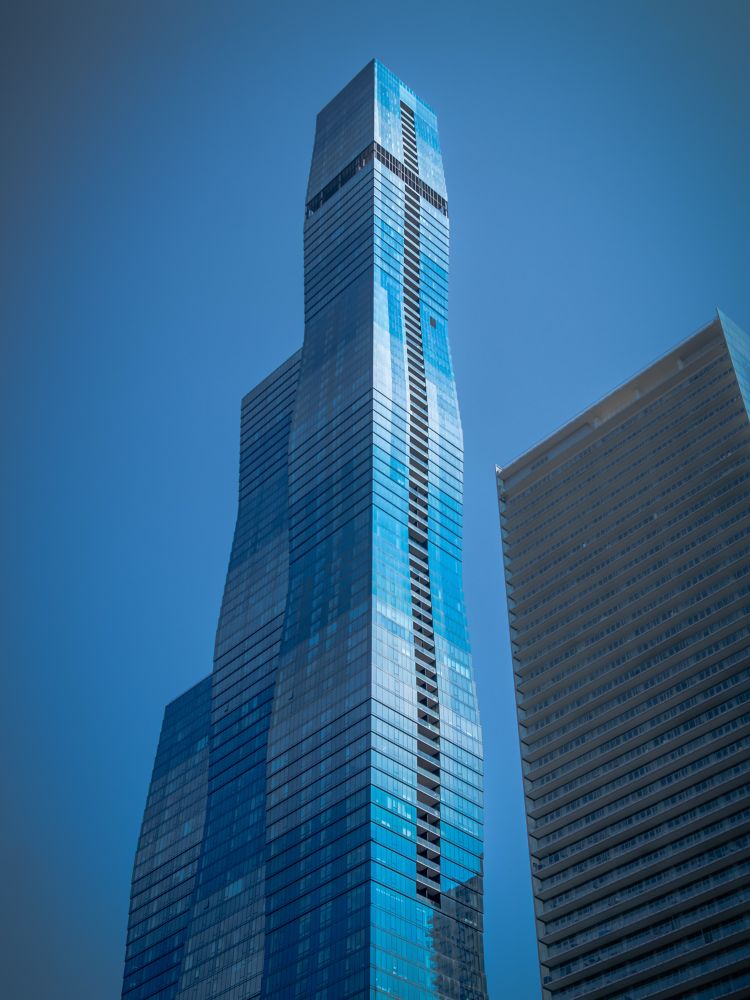 A ground level shot of the Residences at St. Regis along the Chicago River. The wavy tower is multiple shades of blue against a bright blue sky without a cloud to be seen. Another smaller building is in shade on the right side of the frame. 