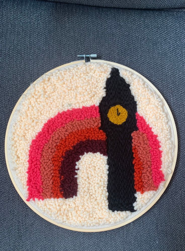 This is a punch needle piece that is 10 inches in diameter. There is a cream colored background with a pink toned rainbow and a black clock tower in the foreground. The hands of the clock tower point to 1:00. Most of the piece is made with looped side of the punch needle giving it a lot of texture, while the clock tower is made with the flat or stitch side of the punch needle. 