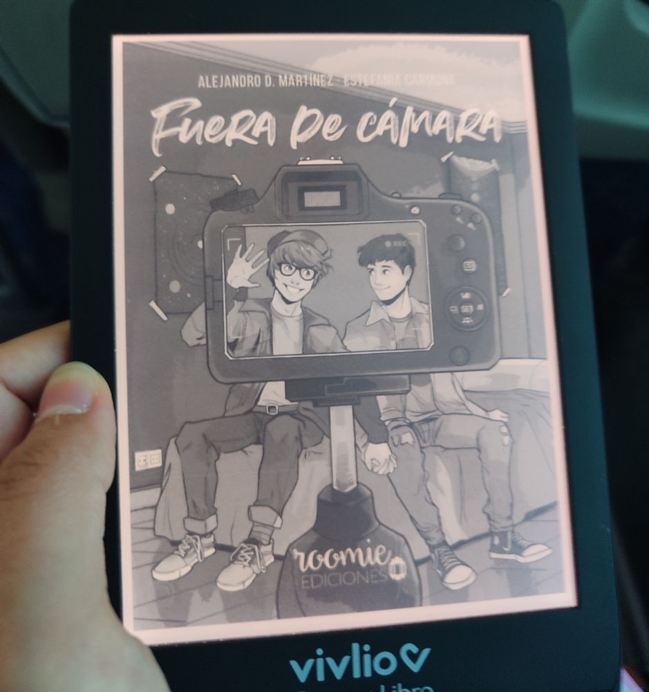Imagen de un lector electrónico marca vivlio (Casa del libro) con la portada del libro "Fuera de cámara" de Alejandro D. Martínez y Estefanía Carmona. En la portada hay dos chicos sonriendo frente a una cámara. Fuera del visor de la cámara se aprecia que están sentados en una cama y se dan la mano.