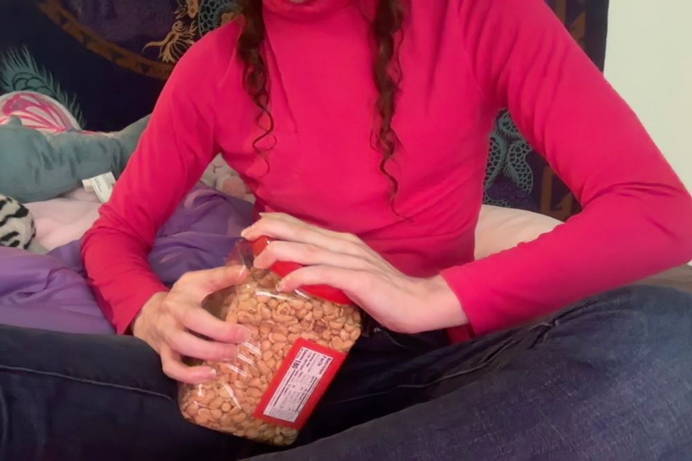 Transgender woman struggles and fails to open Great Value™ peanuts container while sitting on her bed. Blahaj tail visible behind her.