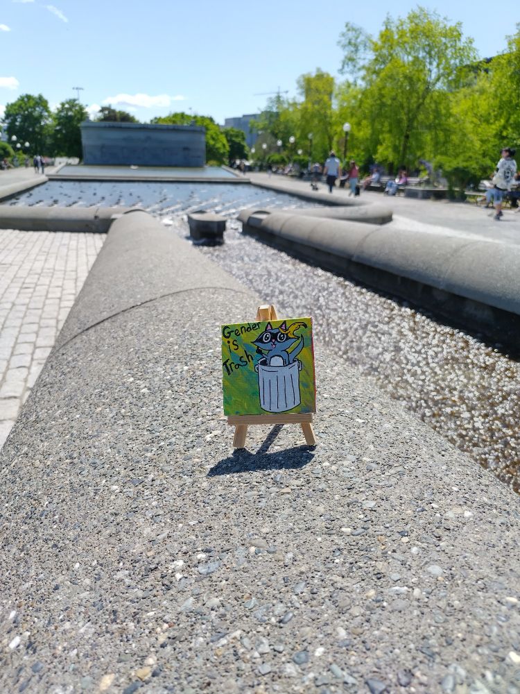 Picture of a miniature painting on a tiny easel resting next to a water fixture at one of the local parks. The picture is of a raccoon in a trash can with a banana peel on its head and next to it says gender is trash. 