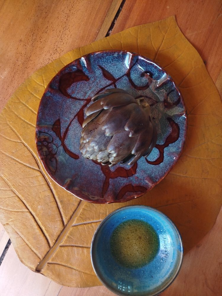 Uma flor de alcachofra com cor mais escura - pois cozida - sobre um prato. Ao lado do prato tem uma tigela pequena com um molhinho. Tudo isso está sobre jogo americano no formato de folha