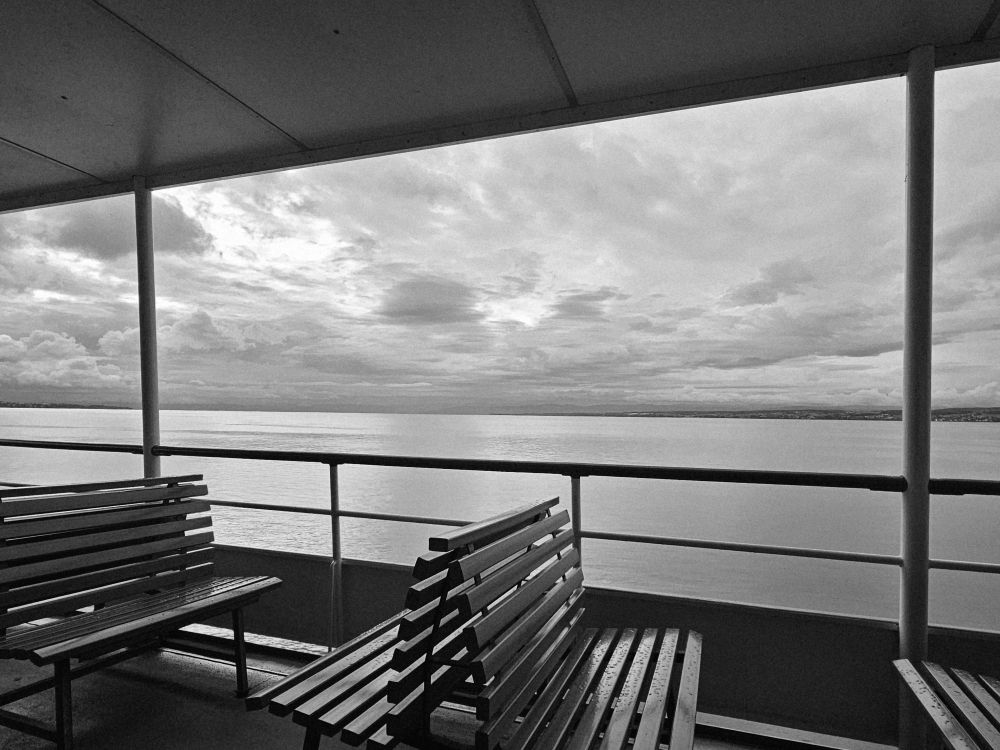 Sitzbänke im Vordergrund, Wasser,Wolkenhimmel. Schwarzweiß. 