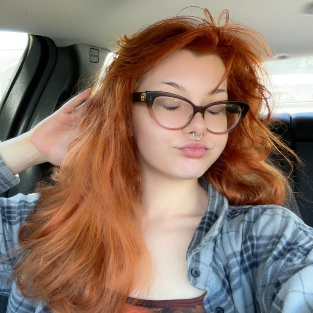 A cute girl sitting in her car, wearing a grey flannel & glasses. The redhead has her eyes closed & is doing a kissy face to the camera. 