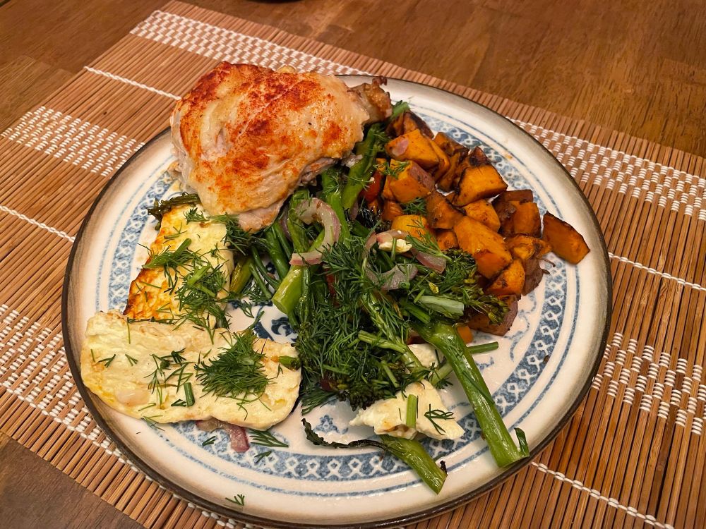 a white plate of food on a bamboo placemat on a wooden table. Chicken, sweet potato, broccolini, and feta garnished with dill. 