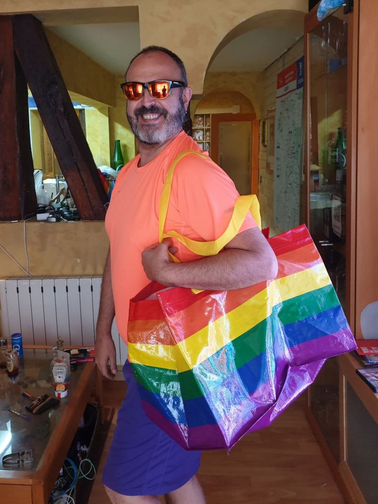 Yo con camiseta naranja y pantalón corto morado llevando al hombro una bolsa Ikea con los colores de la bandera arcoiris.