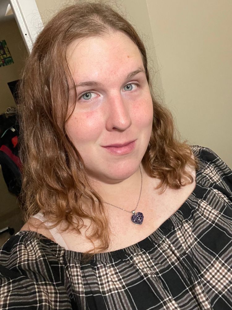A selfie of Lyra bare faced in the bathroom in her brown plaid dress.
