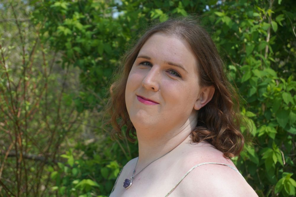 A selfie of Lyra from being three years on HRT this year. She is out in nature with a full face of makeup but not overdone. She has  shoulder length hair again but it is curling and flowing behind her gracefully. She has a purple heart shaped pendant necklace on and you can see the small straps of her green dress. She's turned toward the camera giving a slight smile as she is enjoying herself outside having photos taken of her instead of taking them of herself.