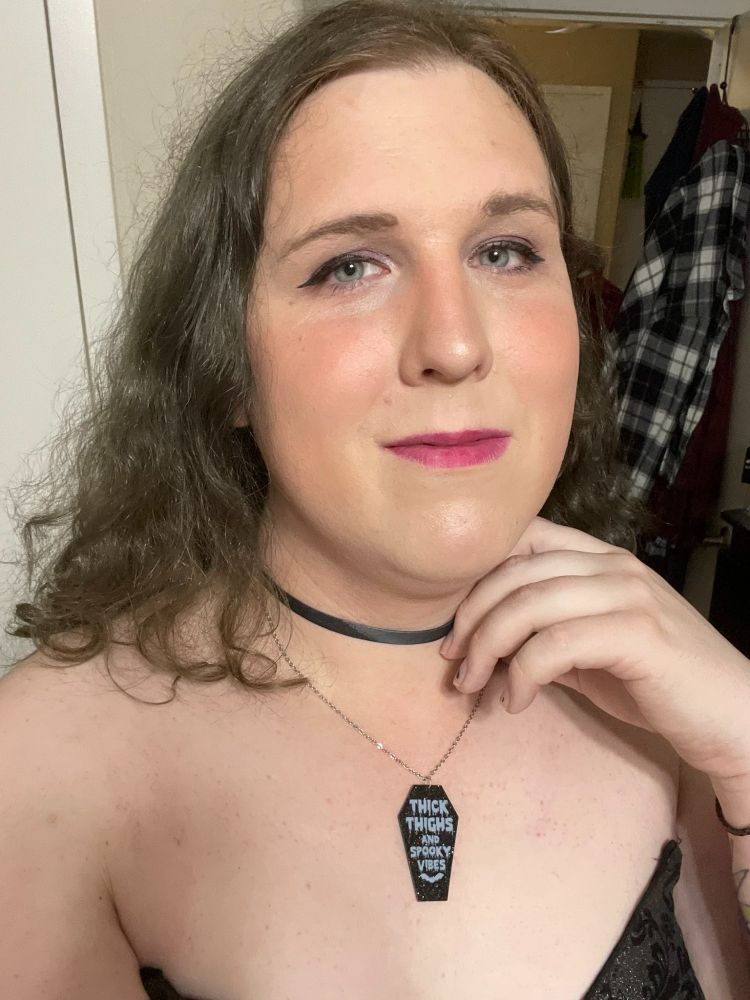 A selfie of Lyra two years into HRT, from last year. She is made up with a full face of makeup that is rather heavy but looks very fem. Her hair is down her back and pouring over her shoulder. She is wearing a black leather choker and a coffin shaped necklace that says "Thick Thighs and Spooky Vibes" on it and a nice black dress that seems to be strapless but it's because one of the straps broke at this point. She is looking at the camera enjoying her newfound beauty.