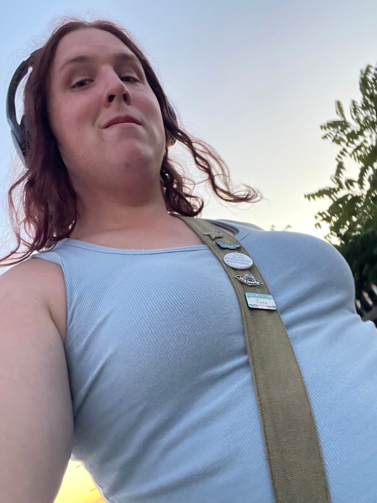 A selfie of Lyra from below smiling in a tank top with their bag strap around them between their boobs making them more prominent along with their sports bra shaping them.