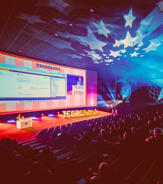 Image taken from the Techorama website. Image shows the main stage with lights and a presentation on the left, illuminated stars on the ceiling, and a darkened audience on the right.