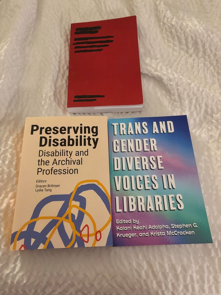 Three books in a pyramid on a white coverlet. The top book has a red cover with blacked out title and authors & is Censorship is a Drag. The bottom left book has an off white color with different color & thickness looping lines at the bottom and a big title that says “Preserving Disability” & small words “Disability & the Archival Profession”. The bottom right book has a blue purple green gradient cover with white letters that say “Trans and Gender Diverse Voices in libraries” & small white letters saying “edited by Keahi Adolpho, Steven G. Krueger, and Krista McCracken”