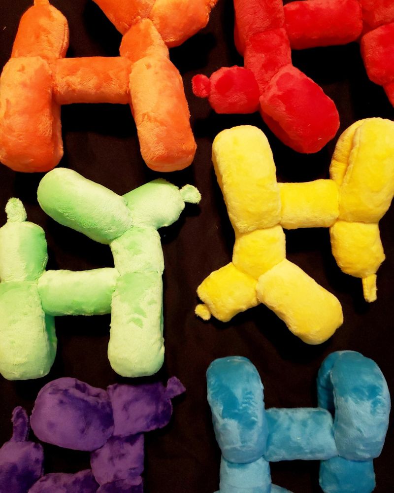 Six plush animals shaped like balloon dogs lined up on a black minky fabric backdrop with two on each row. From top to bottom, there is orange, red, green, yellow, purple, and blue. The balloon dogs on the left side are pointing to the right. The balloon dogs on the right side are upside down, and pointing to the left.