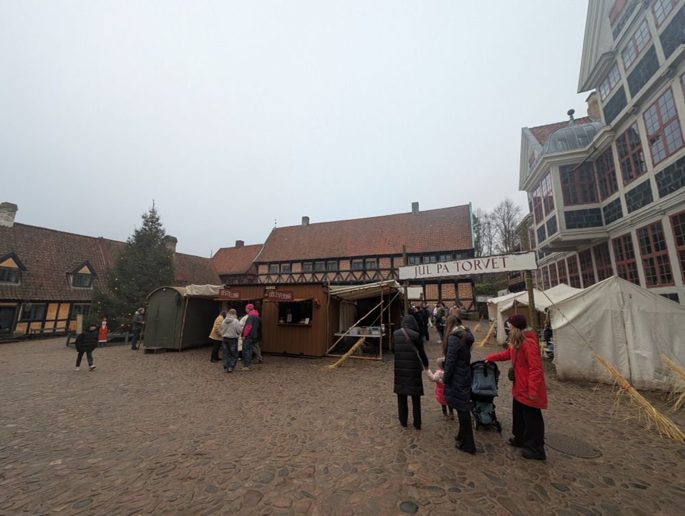 A square in Den Gamle By (The Old Town), Aarhus, Denmark, a sign saying "Jul På Torvet" (Christmas in the Square) is visible, a shop is selling Glogg (Mulled Wine)