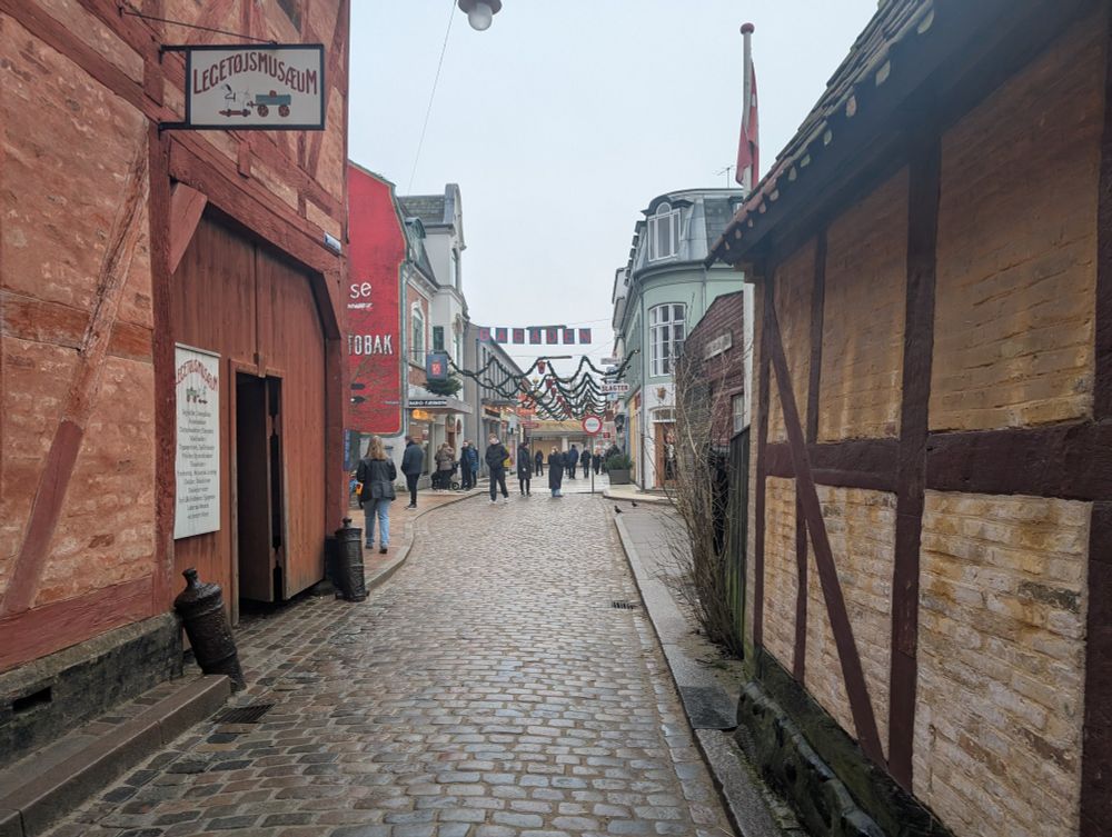 A picture facing down a street in Den Gamle By, Aarhus. A door on the left leads to the Legetøjsmusæum (Toy Museum), ahead is a pedestrian street lined with shops.