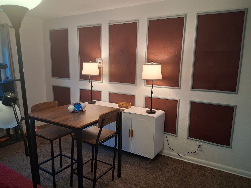Photo of my dining area with the wall panels of burgundy wallpaper and gray molding framing the panels. The sideboard is white with gold-toned handles on the 4 doors. There are 2 stick lamps on the sideboard, one at each end, with a wooden box between them. The table is a high-top with 2 chairs. It is "industrial" style with a wooden top and wrought iron base. The chairs match it.