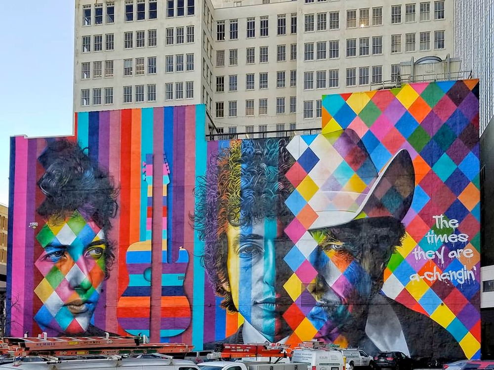 The back of a three-story building with a multi-colored mural of Bob Dylan painted on it. His face is shown at three ages, plus a guitar and the words "the times they are a changin"