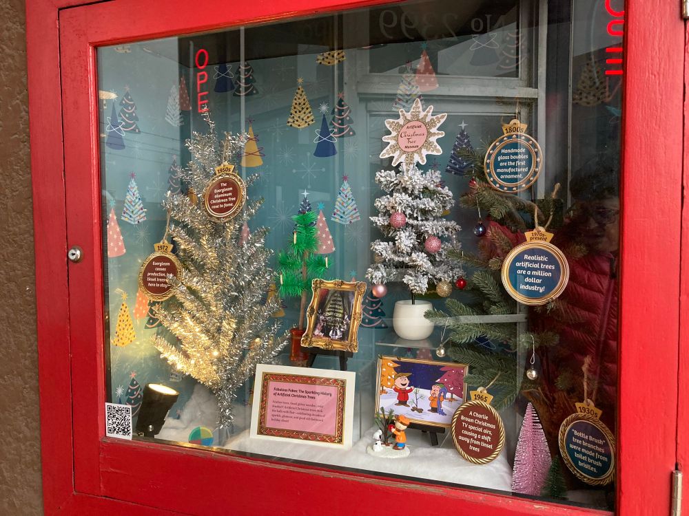 Display of tiny Christmas trees with little explanatory panels inside a glass former firehose box.