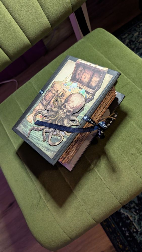 A thick handbound journal sits on a green velvet chair. It has a pirate themed cover and is tied shut with black ribbon