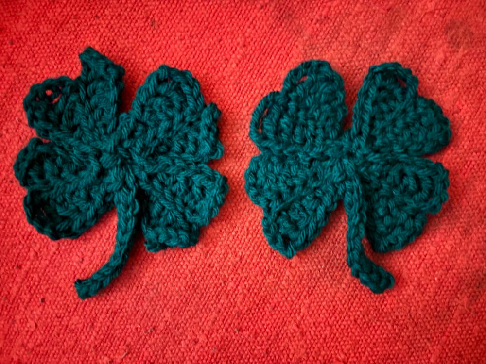 Two crocheted dark green shamrocks, the one on the left is slightly misshapen, the one on the right is more regular shaped 