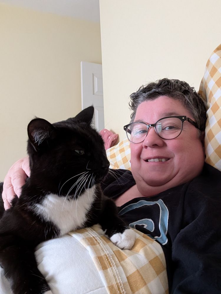 Woman in glasses sitting up in bed. On her chest is a black and white cat. 