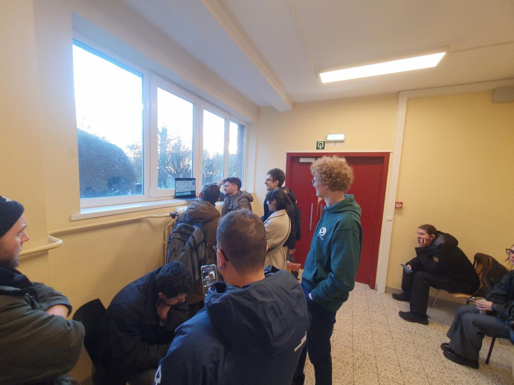 At least 10 people in a room watching a live stream on a laptop.