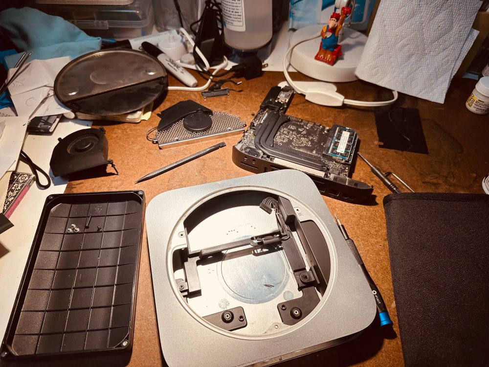 A mid 2011 Mac Mini in pieces on a cluttered drafting table. A small Alf toy from the 90s Wendy’s kids meal standing on a lamp base. 