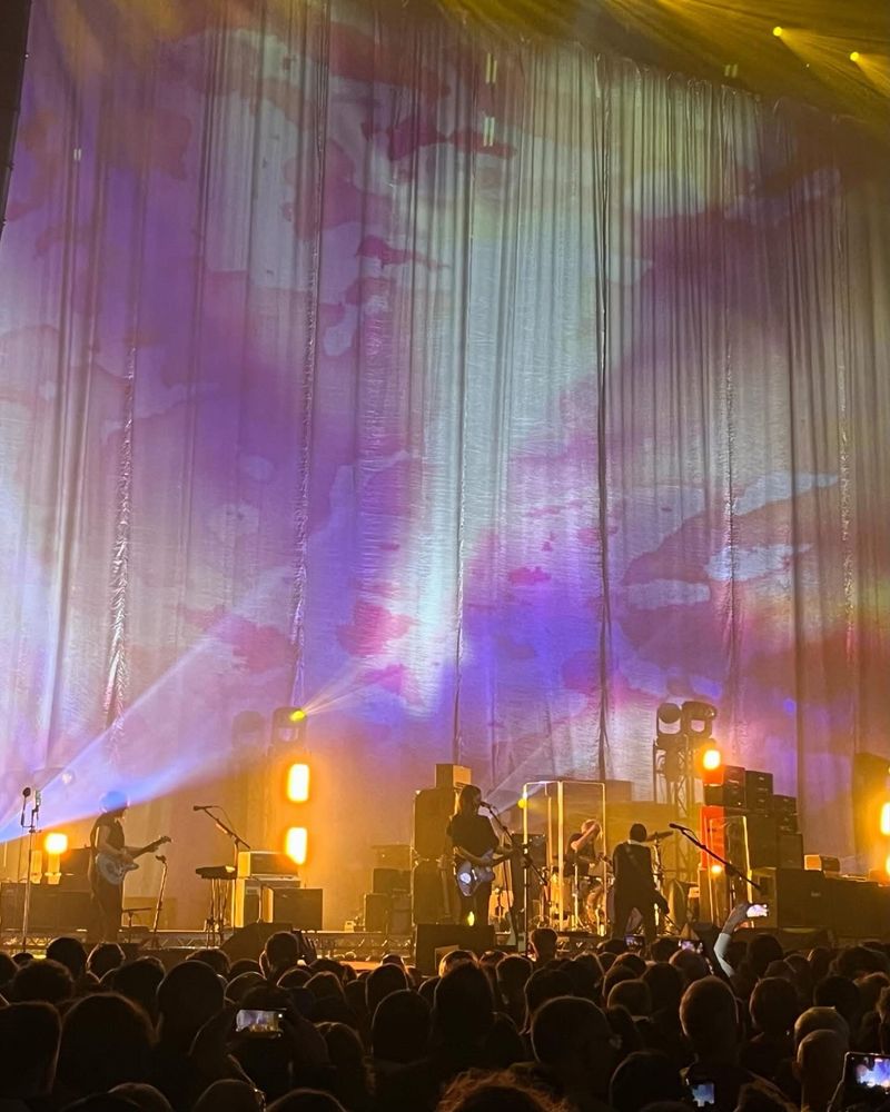 Band on stage from a distance, with a backdrop marbled with pink, lilac, and amber lights. Super shoegazey.